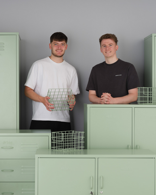 Two men stood behind a group of sage green metal storage lockers and baskets.

